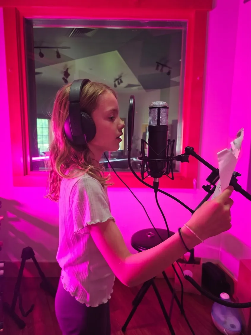 A young singer reading her lyrics at a microphone in a pink-lit recording booth