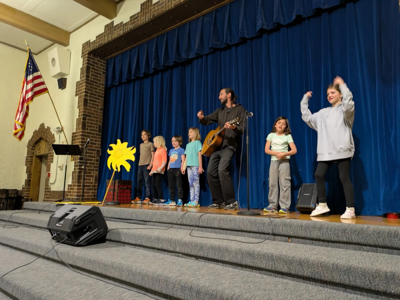 Young students performing on a school auditorium stage while their teacher plays guitar